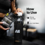Person pouring one scoop of MuscleTech Platinum 100% Creatine Monohydrate powder into a MuscleTech shaker bottle with text instructions showing one serving mixed with 8 ounces of cold water. Demonstrates product use for muscle strength and recovery support.